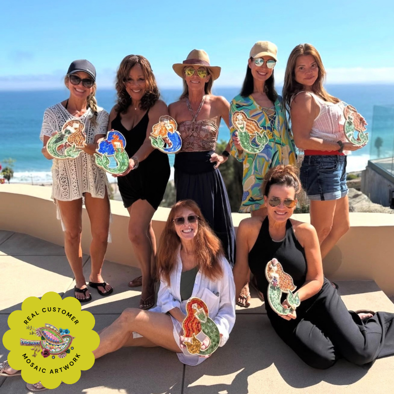 Group of women posing with mermaid mosaic art pieces by the ocean.
